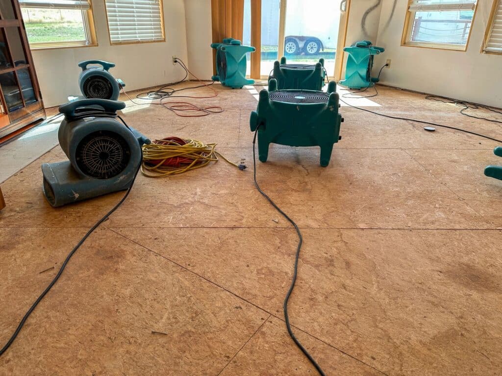 Water damage drying setup with air movers and dehumidifiers placed across a room after floor removal.